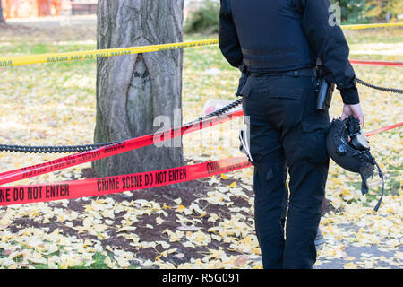 Funzionario di polizia nel casco in piedi dalla scena del crimine chiusa da giallo e rosso nastro cop. Foto Stock