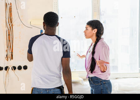 Vista posteriore dell'uomo pittura parete mediante rullo di vernice mentre la sua fidanzata irritata fare spallucce gesto Foto Stock