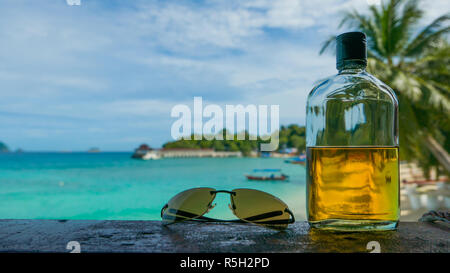 Beach party. Vacanza divertente concetto. Bottiglia di rum e occhiali da sole con Palm tree, la spiaggia, il mare e la barca in background. Foto Stock