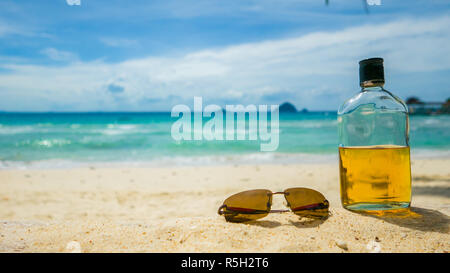 Beach party. Vacanza divertente concetto. Bottiglia di rum e occhiali da sole con spiaggia e mare sullo sfondo. Foto Stock