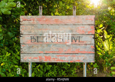 Essere positivo preventivo motivazionale scritta sulla vecchia scheda vintage sign in il forrest, con raggi del sole in background. Foto Stock