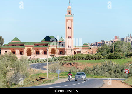 01-03-15, Marrakech, Marocco. Una Moschea e minareto stand come sfondo di un remoto paese road con un auto e scooter guidando lungo di essa nel sub-Atl Foto Stock