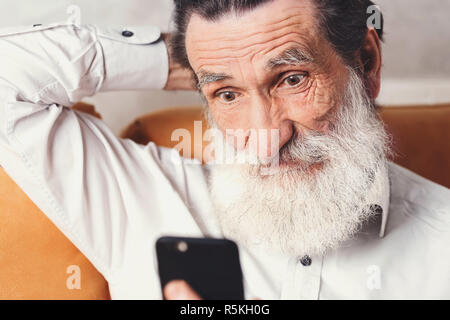 Senior moderno uomo barbuto che indossa una camicia bianca seduto sul divano giallo e utilizza lo smartphone, egli guardando il telefono con anticipazione, egli in attesa nessesary news Foto Stock