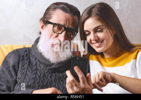 Ritratto di senior padre barbuto in bicchieri considerando lo smartphone mentre è seduto sul divano giallo nel salone luminoso con il suo adulto dai capelli lunghi brunette figlia, tecnologia moderna, concetto di comunicazione Foto Stock