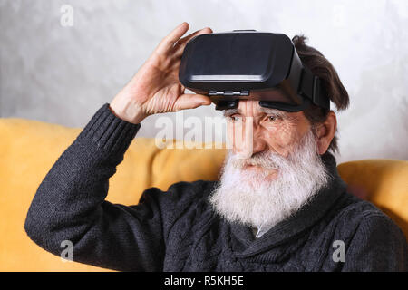 Senior contemporaneo uomo barbuto in grigio pullover in seduta occhiali VR sul divano giallo nella luce soggiorno, la tecnologia del futuro concetto Foto Stock