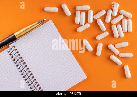 Le capsule di medici e pillole intorno a un vuoto portatile con penna su uno sfondo arancione, close-up, vista dall'alto, con copia spazio. Foto Stock