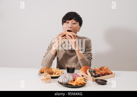 Ritratto di uomo asiatico seduto e snacking ciambelle isolate su sfondo bianco. Uomo grasso concetto di dieta Foto Stock