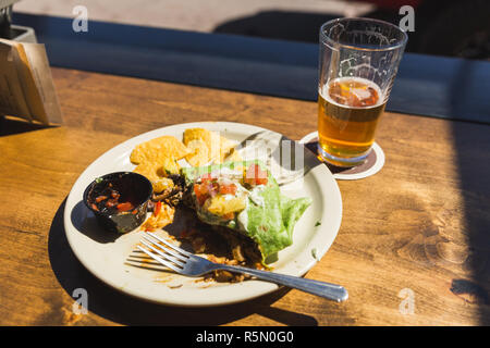Lastra di vetro su un tavolo di legno con burrito, patatine, salsa e un semi-bevuto una pinta di birra. Esterno l'impostazione diurna. Foto Stock