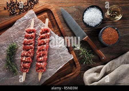 Materie Turkish Urfa kebab sul bianco di carta di cottura e il tagliere di legno. Decorate con erbe aromatiche, spezie e Coltello per cuoco. Vista aerea. Foto Stock
