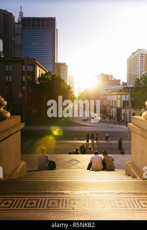 La gente seduta sulle scale di casa del Parlamento la visione di Melbourne henge, quando il sole tramonta in linea con la griglia del CBD di Melbourne, Australia Foto Stock