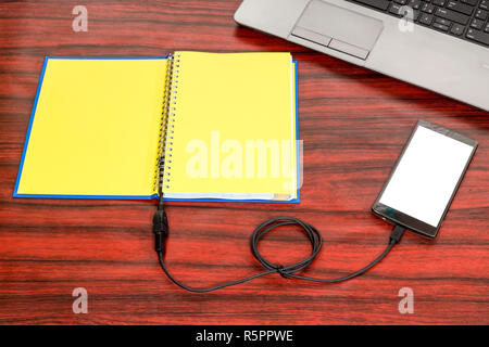 Aprire un libro di testo collegato a un telefono cellulare tramite cavo USB, posa accanto a un computer portatile e matita su una scrivania. Foto Stock