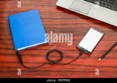Prenota collegato ad un telefono cellulare tramite cavo USB, posa accanto a un computer portatile e matita su una scrivania. Foto Stock