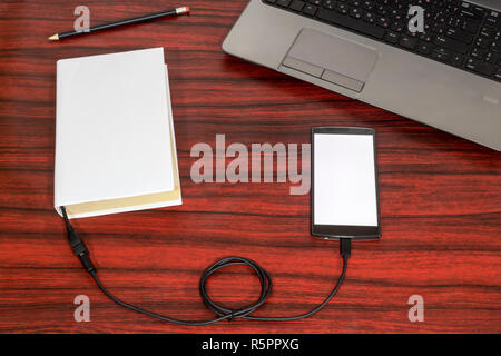 Prenota collegato ad un telefono cellulare tramite cavo USB, posa accanto a un computer portatile e matita su una scrivania. Foto Stock