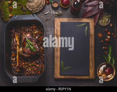 Slow cucinato Arrosto di capriolo in ghisa nera pan, vista dall'alto. Brasato di gamba di cervo con osso e verdure arrosto in pentola su dark cucina rustica tabella b Foto Stock