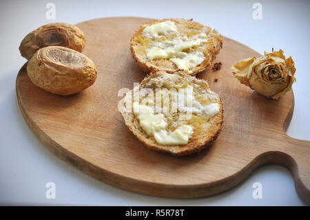 Due spigoli delle fette di pane con burro, due patate al forno e un bianco secco rose decorazione su una tavola di legno Chopboard. Vista ravvicinata della colazione casalinga Foto Stock