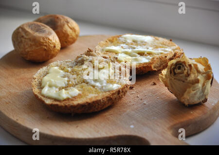 Due spigoli delle fette di pane con burro, due patate al forno e un bianco secco rose decorazione su una tavola di legno Chopboard. Vista ravvicinata della colazione casalinga Foto Stock