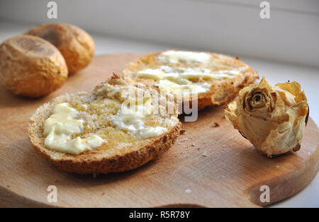 Due spigoli delle fette di pane con burro, due patate al forno e un bianco secco rose decorazione su una tavola di legno Chopboard. Vista ravvicinata della colazione casalinga Foto Stock