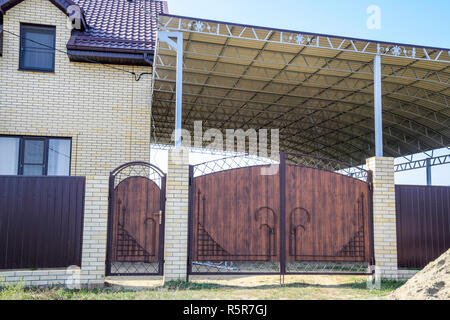 Casa di mattoni con una recinzione e cancelli. Una grande tenda con una cornice di acciaio. Vista di una nuova built-up recinzione e una casa fatta di mattoni e lamiera ondulata. Foto Stock