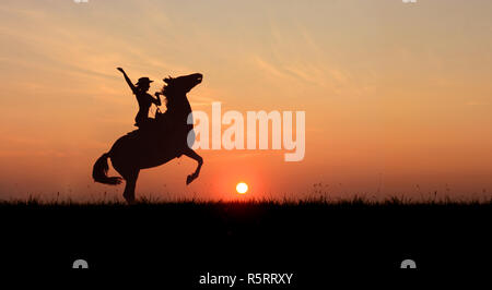 Cowgirl in sella ad un cavallo, allevamento fino al tramonto. Stallone in piedi sulle zampe posteriori in linea di orizzonte con il sole al tramonto. Foto Stock
