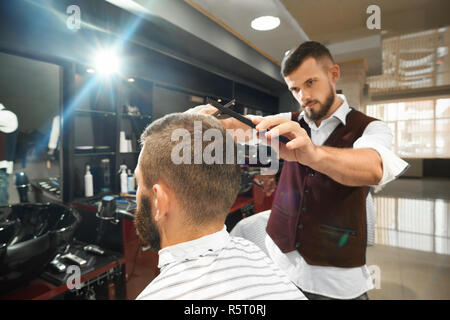 Grave brutale barbiere in uniforme in piedi vicino a client maschile seduto in poltrona e facendo elegante taglio di capelli in barberia. Qualificato mediante l uomo e il pettine rasoio. Concetto di processo di lavoro. Foto Stock