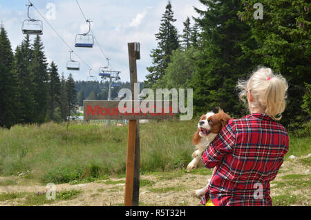 Donna che mantiene il suo cane - Cavalier King Charles Spaniel - in piedi dal cartello in legno per il carrello di montagna in colline Foto Stock