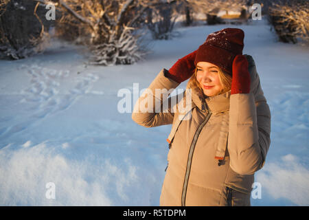 Piuttosto giovane donna caucasica in rosso mezzoguanti e red winter hat godendo di buona soleggiata giornata invernale in presenza di neve la natura. Lei tiene il suo cappello e cercando di sun. Buon umore, meteo e in inverno il concetto di amore. Foto Stock