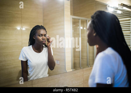 Focalizzato bella giovane donna pulizia la sua pelle con un tampone di cotone, guardando lo specchio nel bagno di casa. Bellezza e cura della pelle concetto, lo stile di vita Foto Stock