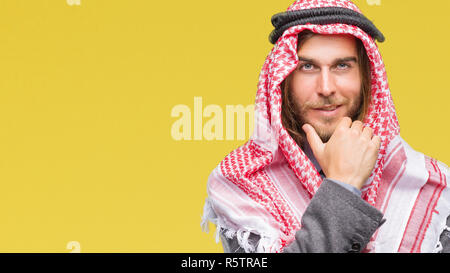 Giovane bello arabian uomo con capelli lunghi che indossa keffiyeh su sfondo isolato guardando fiduciosi alla fotocamera con sorriso con bracci incrociati e h Foto Stock