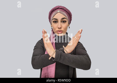 Grave giovane donna araba di alzarsi e di tenere le mani in posizione crpss. Lei si guarda sulla fotocamera. Modello indossa hijab e abito nero. Isolato su backgroun grigio Foto Stock