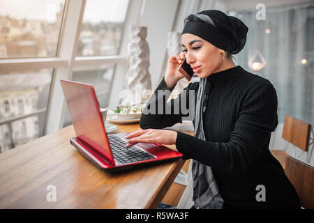 Occupato giovane donna araba sedersi a tavola all'interno. Ella parla al telefono e di lavoro sul computer portatile. Modello guarda verso il basso Foto Stock