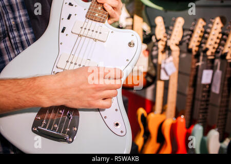 Close up uomo con luce blu-chitarra in mani. Egli svolge su di esso e tocca la sua stringhe. Molte colorate chitarre elettronico appeso alla parete dietro Foto Stock