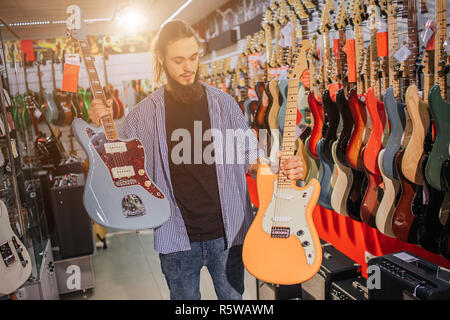 Giovane uomo tenere due coloratissimi chitarre elettriche. Egli guarda uno giallo. Molte altre chitarre elettriche sono dietro di lui. Egli stand alone in msuic shop Foto Stock