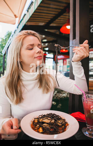 Giovane bella donna mangiare pasta nera con piatti a base di frutti di mare e nero di seppia nel ristorante all'aperto. Divertente e bello. Foto Stock