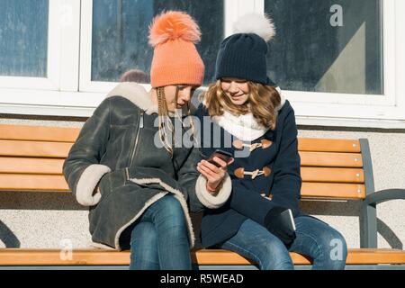 Close-up esterna inverno ritratto di due ragazze adolescenti studenti nel profilo sorridente e parlando, ragazze guardando al telefono cellulare. Foto Stock