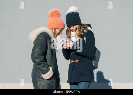 Close-up esterna inverno ritratto di due ragazze adolescenti studenti nel profilo sorridente e parlando, ragazze guardando al telefono cellulare. Foto Stock
