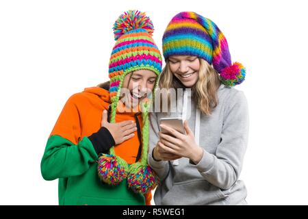 Inverno ritratto di due happy belle ragazze adolescenti in maglia cappelli divertendosi con telefono cellulare, isolato su sfondo bianco. Persone, giovani un Foto Stock