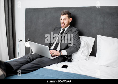 Imprenditore elegante in tuta a lavorare con il computer portatile sul letto della camera in hotel durante un viaggio di lavoro Foto Stock