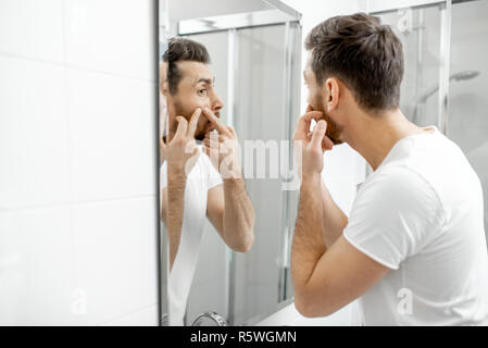 Uomo in t-shirt bianca con la spremitura di acne sul viso cercando di lo specchio del bagno Foto Stock