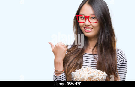 Giovane donna asiatica mangiare popcorn su sfondo isolato di puntamento e mostra con il pollice fino a che il lato con buon viso sorridente Foto Stock