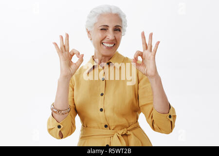 Carismatica, felice ed eccitato carino madre anziana con i capelli grigi in giallo trendy trench coat che mostra bene i gesti e baluginare in omologazione gradimento Foto Stock