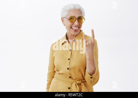 Il suo anime è giovane. Ritratto di gioiosa lieto e spensierato entusiasta nonna con i capelli grigi in Eleganti occhiali da sole e rivestimento giallo sporgente Foto Stock