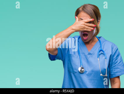La mezza età senior medico infermiere donna sullo sfondo isolato peeking in stato di shock che copre il viso e gli occhi con la mano, guardando attraverso le dita con embarras Foto Stock
