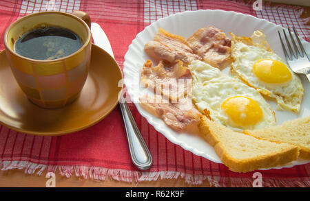 La deliziosa prima colazione - una tazza di caffè, un piatto di uova fritte, pancetta e toast, accanto alle posate sul rosso a scacchi igienico Foto Stock