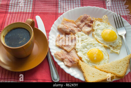 La deliziosa prima colazione - una tazza di caffè, un piatto di uova fritte, pancetta e toast, accanto alle posate sul rosso a scacchi igienico Foto Stock