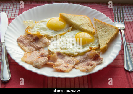 Colazione gustoso piatto di uova fritte, pancetta e toast, accanto alle posate sul rosso a scacchi igienico Foto Stock