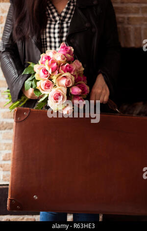 Valigia vintage e la ragazza con un mazzo di rose. Fiori e accessori per viaggiare in prossimità della parete di mattoni. Foto retrò. Spazio libero per copiare il testo in Foto Stock
