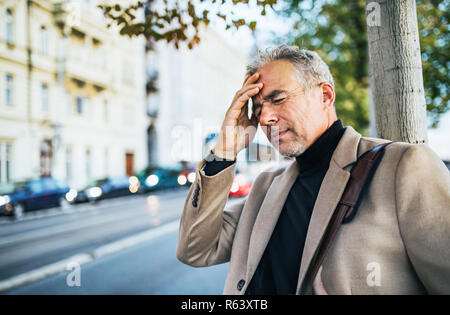 Imprenditore maturo nel dolore in piedi su una strada in città, tenendo la fronte. Foto Stock