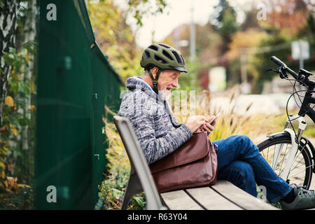 Un uomo anziano con electrobike seduta su una panchina all'aperto in città, utilizza lo smartphone. Foto Stock