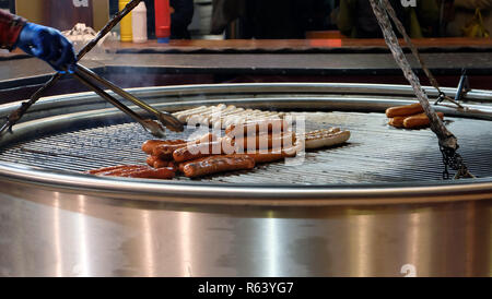 Salsicce tedesche o wurst essendo alla griglia su un grande griller in metallo. Una mano con pinze da cucina può essere visto il ribaltamento della salsiccia. Foto Stock