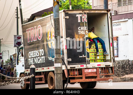 Un uomo dorme su casse di birra "Trio" nel cassone aperto di un camion che attraversa Freetown, Sierra Leone, con pedoni e un agente di polizia in piedi sul ciglio della strada. Foto Stock